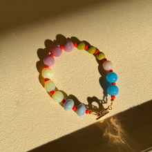 Load image into Gallery viewer, A beaded bracelet with pastel-colored round beads in pink, yellow, blue, green, and white, separated by small red spacer beads, featuring a gold clasp, displayed on a textured beige surface under direct sunlight.
